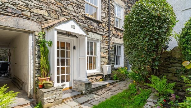 A stone building with a white door, a small porch, and a garden with various plants. - 2 Bed in Ambleside (oc-lr166) (Ambleside)