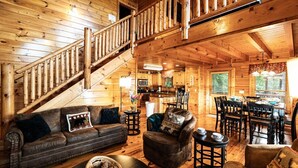 Interior de una cabaña de madera con escalera, una sala de estar con sofá y sillones, un comedor y una cocina.
