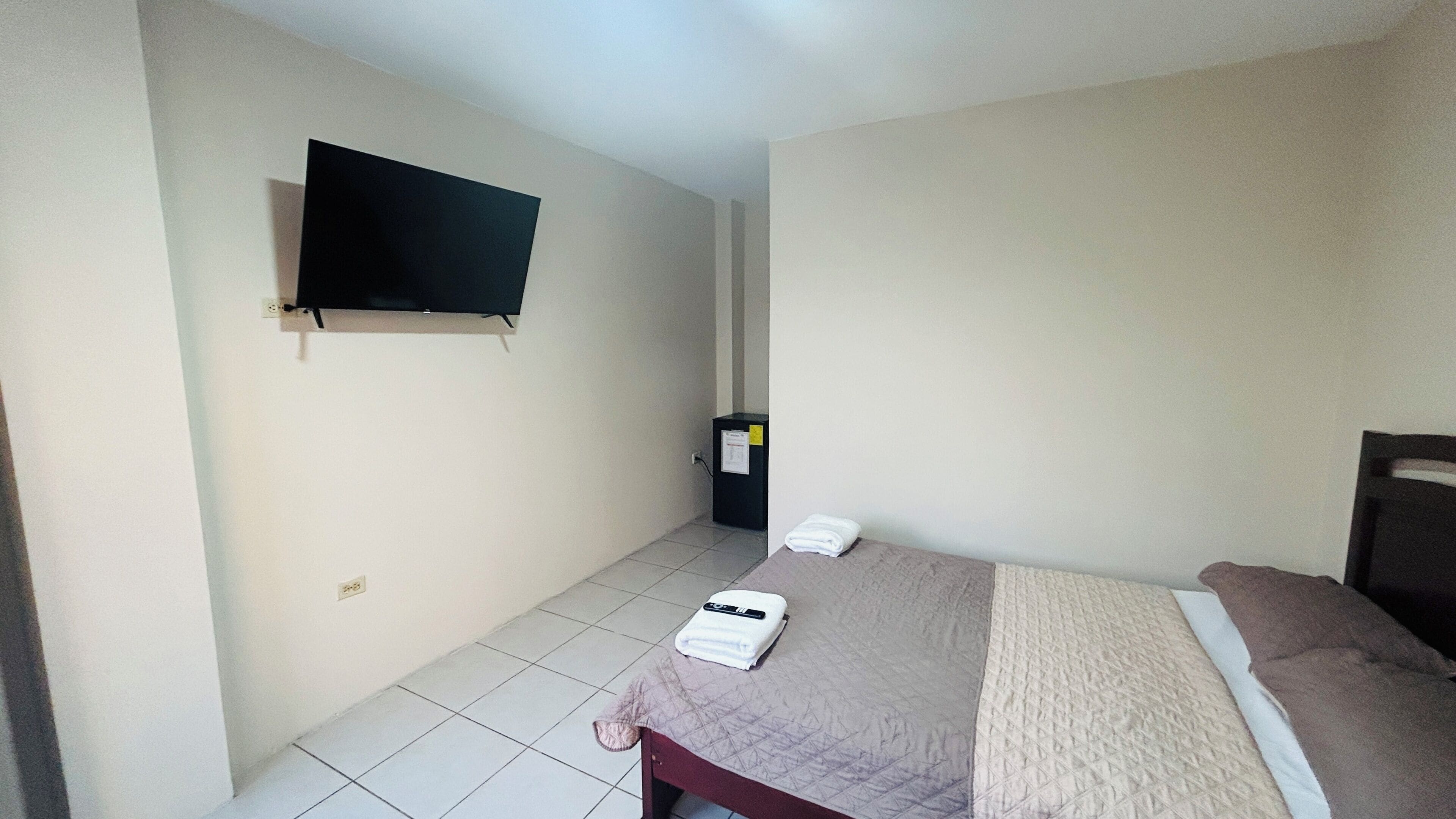 A hotel room with a bed, a television mounted on the wall, a small cabinet, and a towel on the bed.