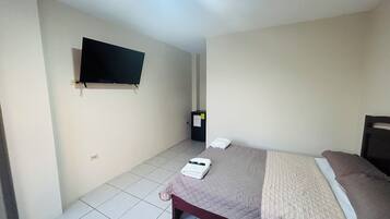 A hotel room with a bed, a television mounted on the wall, a small cabinet, and a towel on the bed.