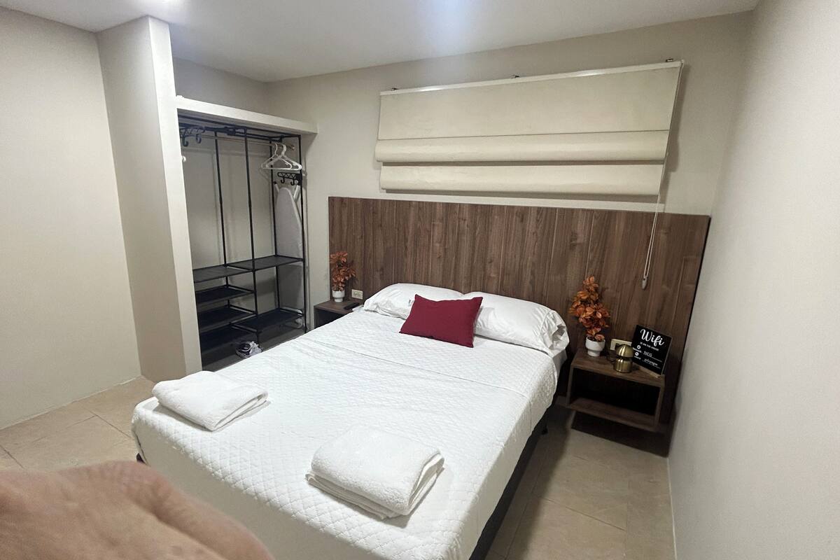 A hotel room with a bed, a wooden headboard, a nightstand, a towel, a hanging shower curtain, and a sign indicating WiFi availability.