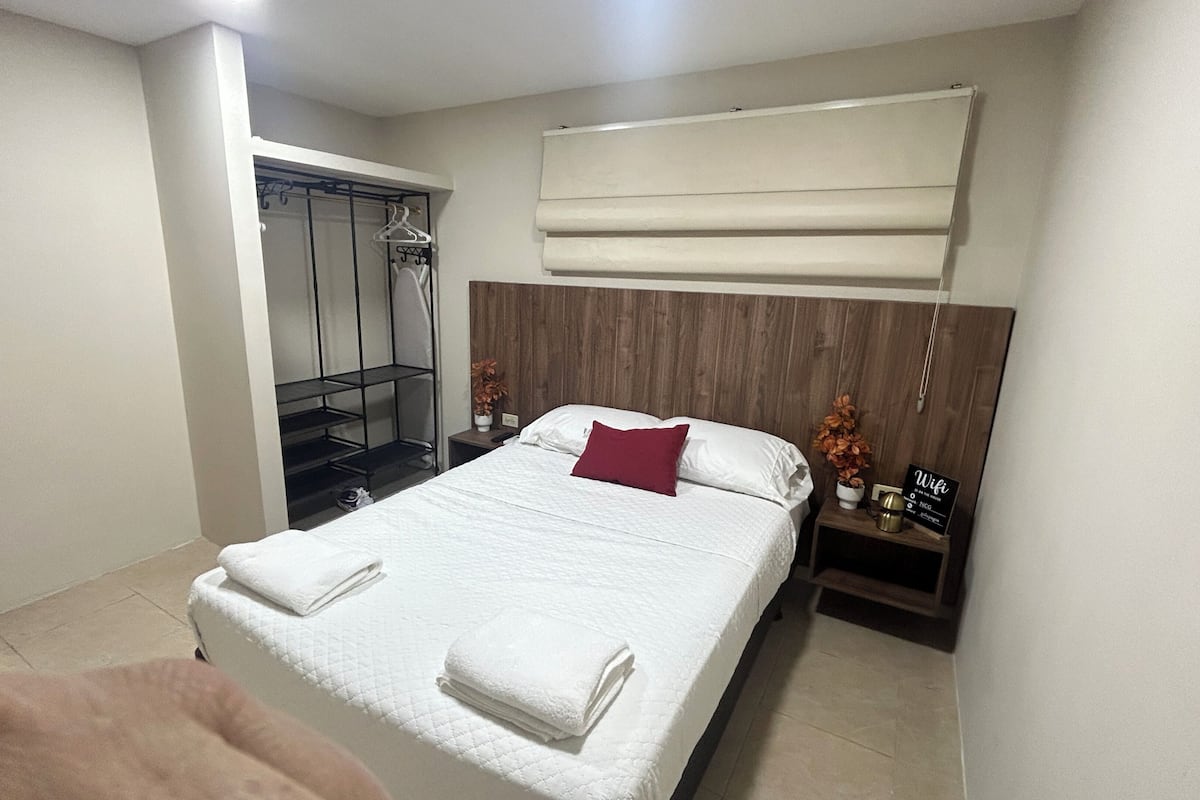 A hotel room with a bed, a wooden headboard, a nightstand, a towel, a hanging shower curtain, and a sign indicating WiFi availability.