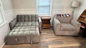 Une pièce comprenant un canapé d’angle, une petite banquette, une table de chevet en bois, une lampe et une fenêtre avec des stores.