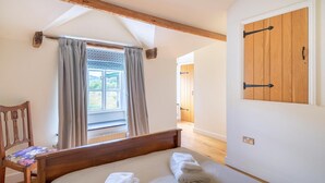 Ein Schlafzimmer mit einem hölzernen Bett, einem fenster mit Vorhängen, einem Holzschrank und einem Sessel mit floralem Kissen.