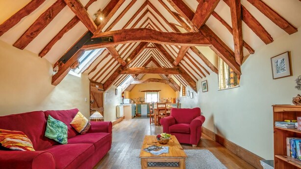 Ein geräumiges Wohnzimmer mit hoher Decke, Holzbalken, einem roten Sofa, einem Holztisch und einem Bücherregal.