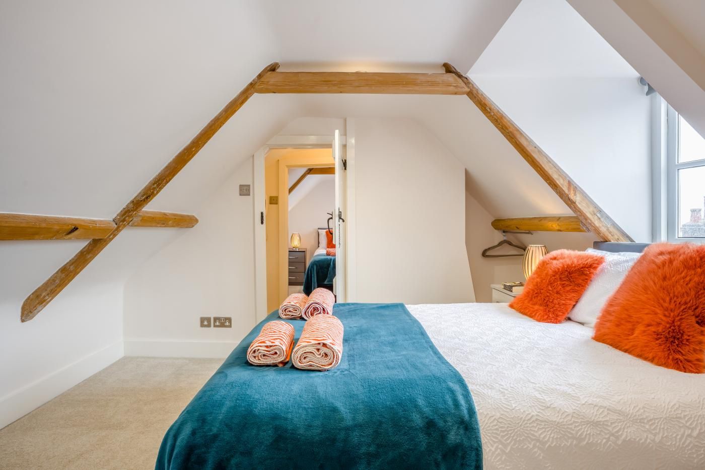 A bedroom with a sloped ceiling, a bed with a teal bedspread and orange pillows, and a view through an open door.