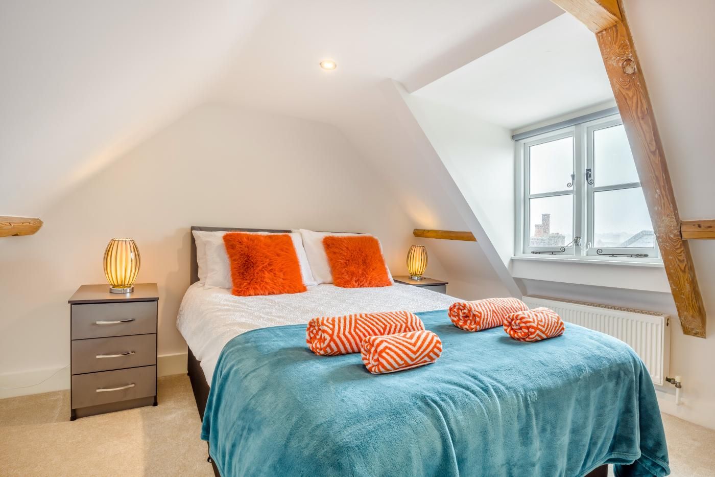 A bedroom with a sloped ceiling, a bed with orange pillows, a blue blanket, and two bedside lamps.