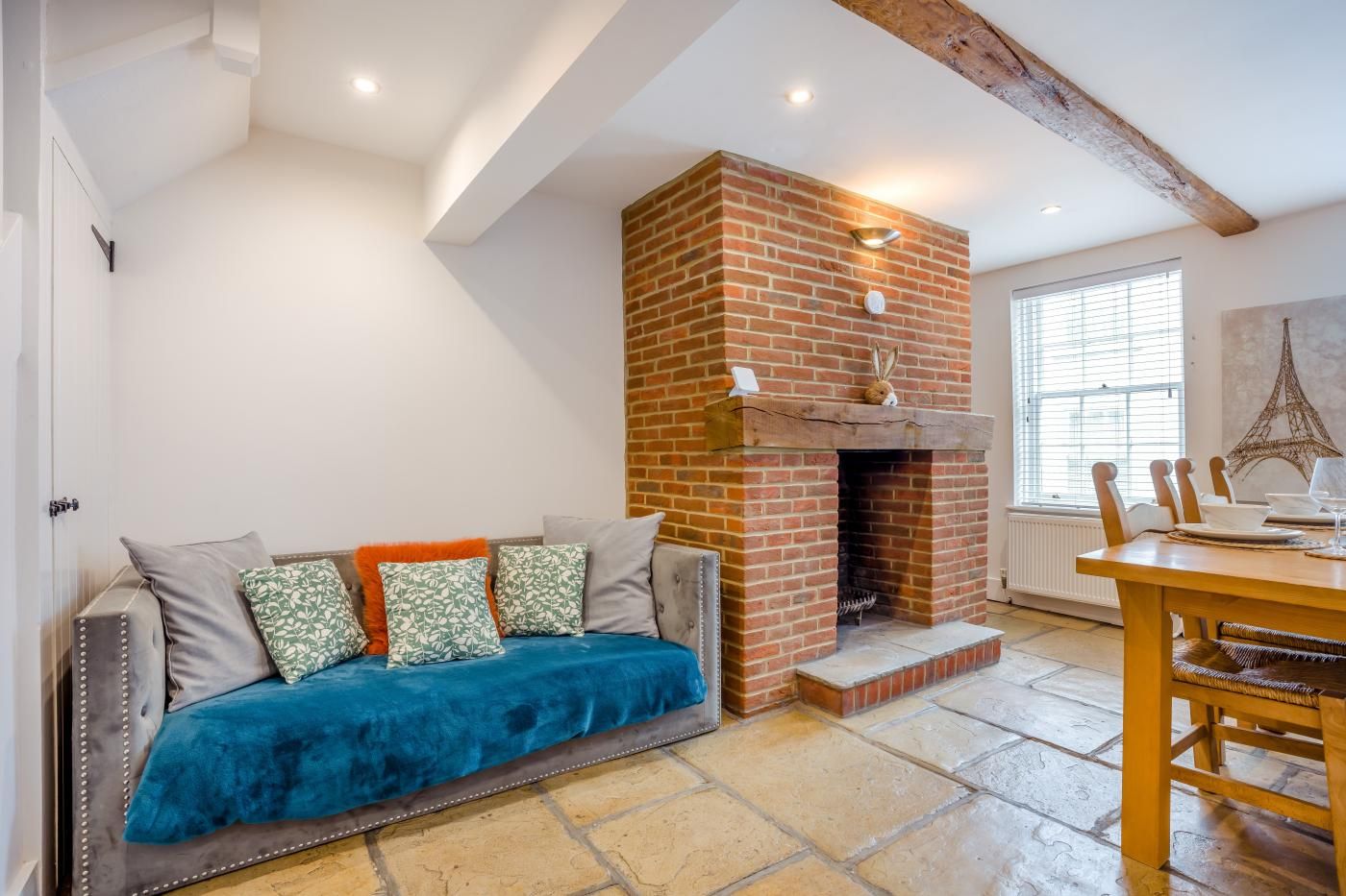 A cozy living room with a brick fireplace, a sofa with cushions, a dining table with chairs, and a large window with blinds.