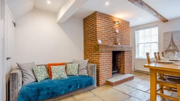 A cozy living room with a brick fireplace, a sofa with cushions, a dining table with chairs, and a large window with blinds.