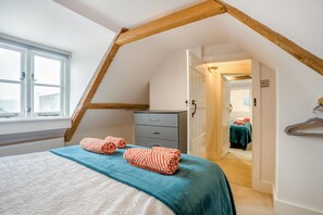 Ein Schlafzimmer mit schrägen Wänden, einem Bett mit türkisblau gestreifter Bettdecke, zwei gestreifte Handtücher, eine Kommode und eine Tür zu einem anderen Raum.