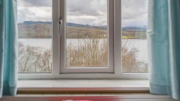Una stanza con un letto, una finestra con tende e vista su un lago e delle colline.