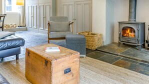 A cozy living room with a stone fireplace, a leather sofa, a grey armchair, and a wooden chest of drawers. - 3 Bed in Skelwith Bridge (oc-lr033) (Skelwith)