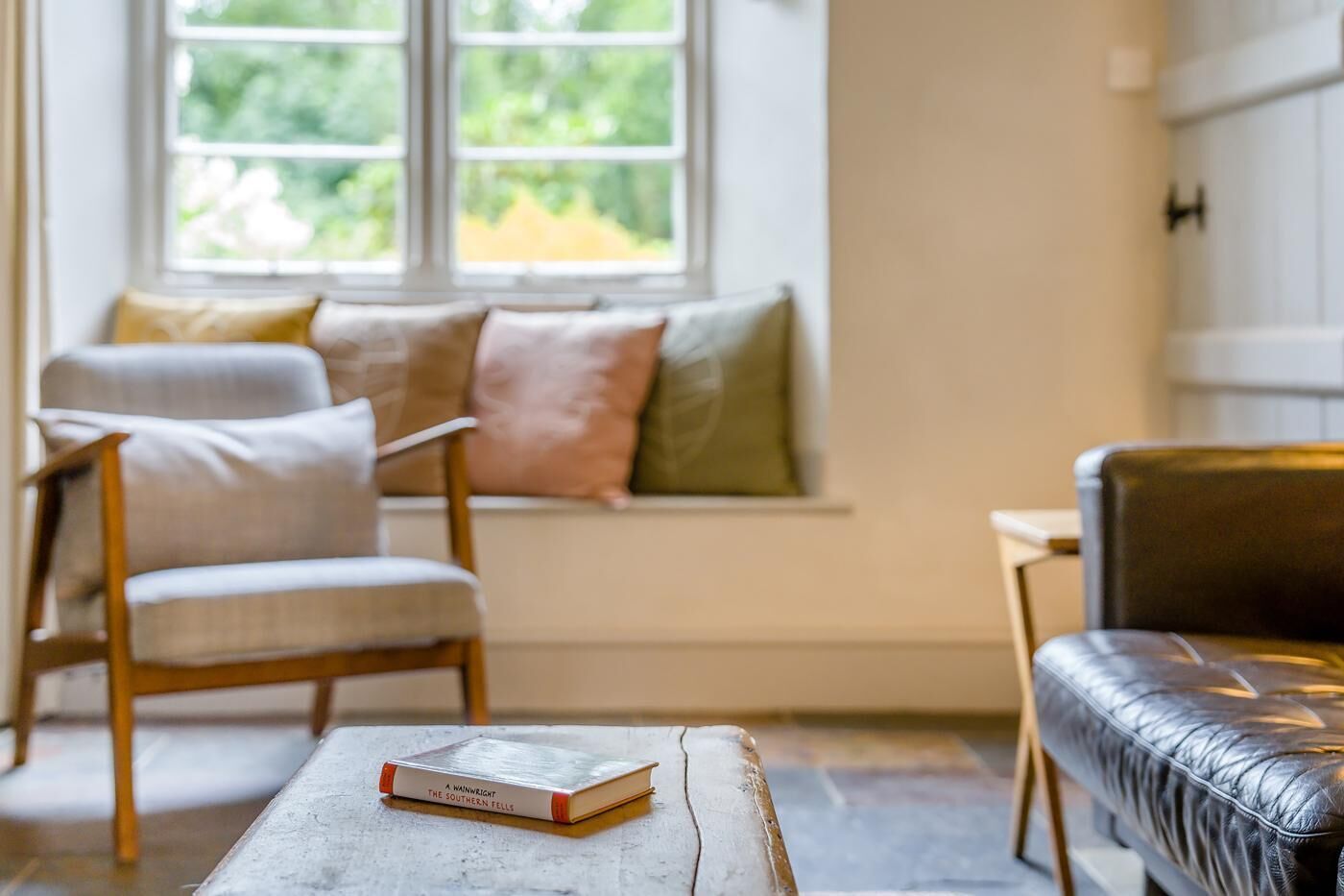 A cozy seating area with a wooden chair and a cushioned bench, a book on a small table, and a window with a view of greenery.