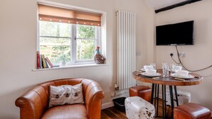 Ett rum med en brun lädersoffa, ett runt träbord med vita tallrikar och glas, ett vitt element, ett fönster med persienner och en väggmonterad TV.