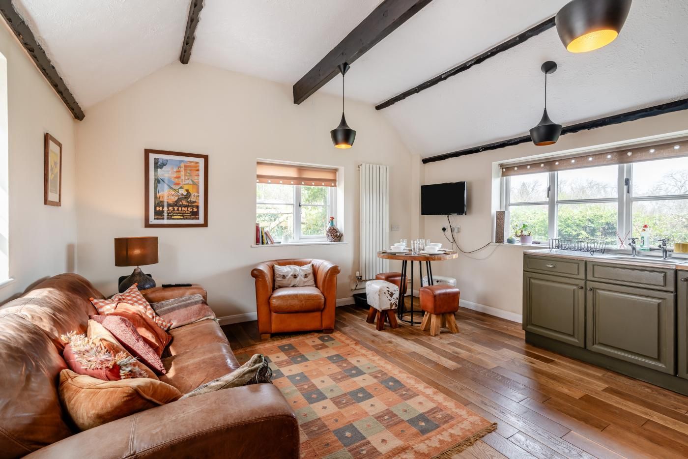 A cozy living room with a leather sofa, a patterned rug, a wooden dining table with chairs, a kitchen area with green cabinets, and a large window with a view of greenery.