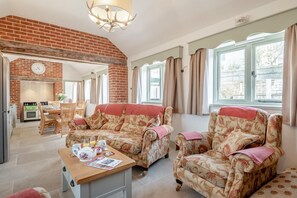 Ein Wohnzimmer mit Kamin aus Ziegelsteinen, zwei gemusterten Sesseln, einem Holztisch und einem Essbereich mit Tisch und Stühlen.
