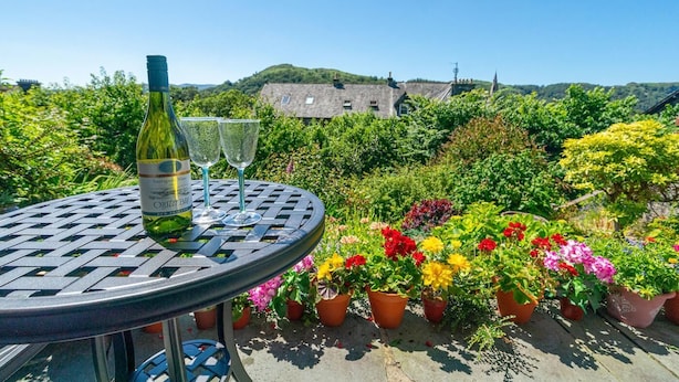 A round metal table with a bottle and two glasses, surrounded by colorful potted plants and a stone building in the background. - 1 Bed in Ambleside (oc-lr077) (Ambleside)