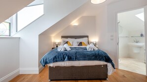 Quarto com teto inclinado, cama com cobertura azul, luminárias de cabeceira e vista para um banheiro.
