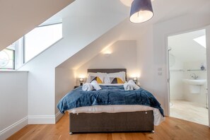 Ein Schlafzimmer mit schrägen Wänden, einem Bett mit blauer Bettdecke, Nachttischlampen und Blick ins Bad.