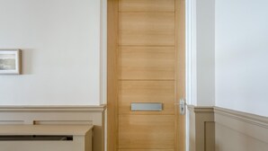 Een houten deur met een handgreep en een klein rechthoekig plaatje, een witte muur met een fotolijst en een beigekleurde plint.