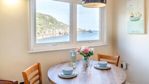 A dining area with a table, chairs, a vase of flowers, and a view of the sea through a window. - 1 Bed in Lamorna Cove, (oc-h40239) (Lamorna)