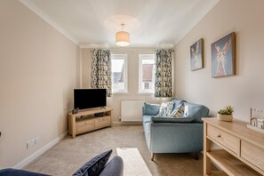 A living room with a television on a wooden stand, a blue sofa, a wooden cabinet, and a window with patterned curtains. - 2 Bed in Kelso (oc-w39065) (Kelso)