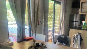 Un coin repas avec une table en bois, des chaises blanches, un vase contenant des plantes séchées et une vue par la fenêtre.