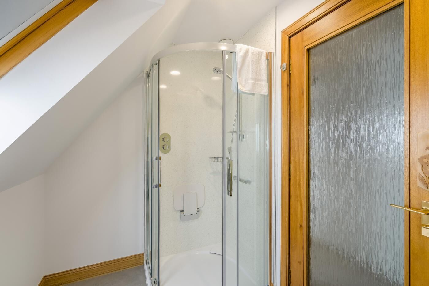 Une douche fermée par des parois de verre, avec une serviette accrochée à la porte, un cadre de porte en bois et un plafond blanc.
