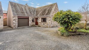 A stone building with two garage doors, a gravel driveway, and a small garden with a tree. - 3 Bed in Pitlochry (oc-t37864) (Pitlochry)