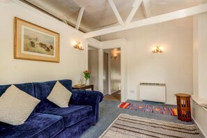 A living room with a blue sofa, a framed picture, wall-mounted lights, a radiator, and a patterned rug. - 1 Bed in Upton Cross (oc-c34217) (Caradon Town)
