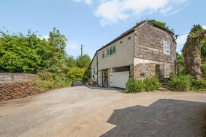 A two-story stone house with a garage, surrounded by greenery and a gravel driveway.