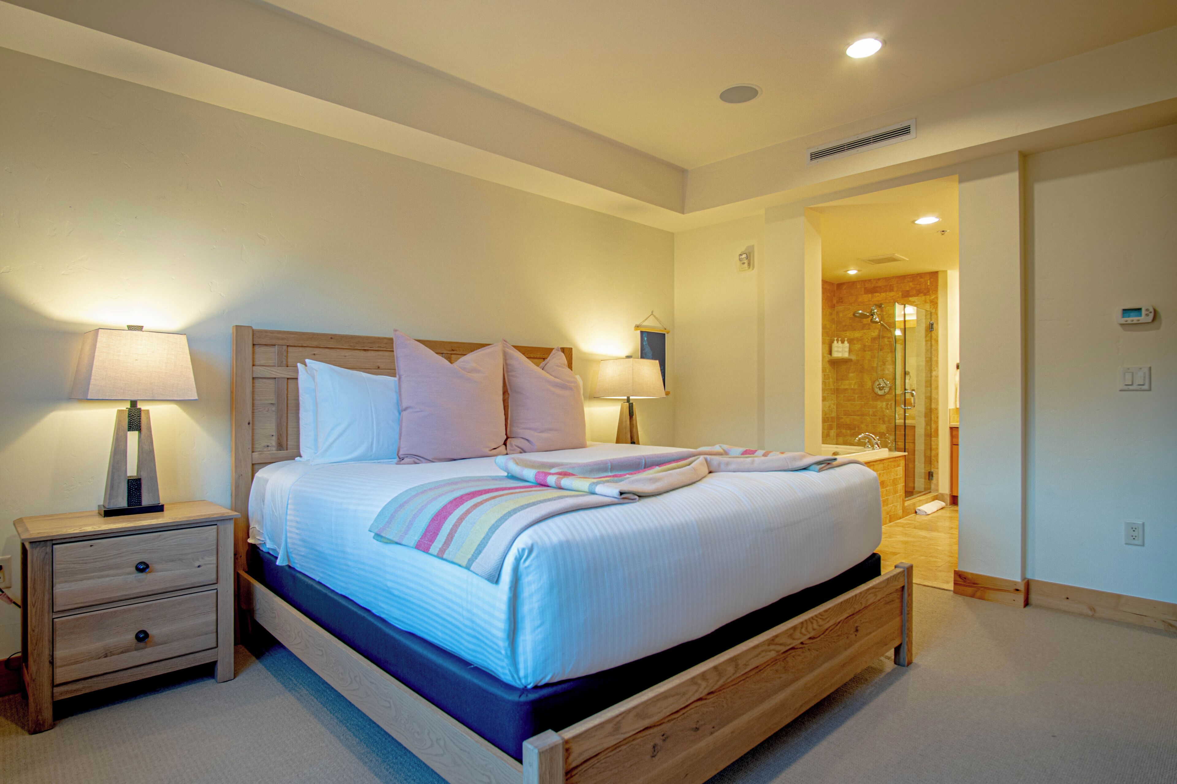 A bedroom with a bed, bedside tables, a lamp, and a bathroom visible through an open door.
