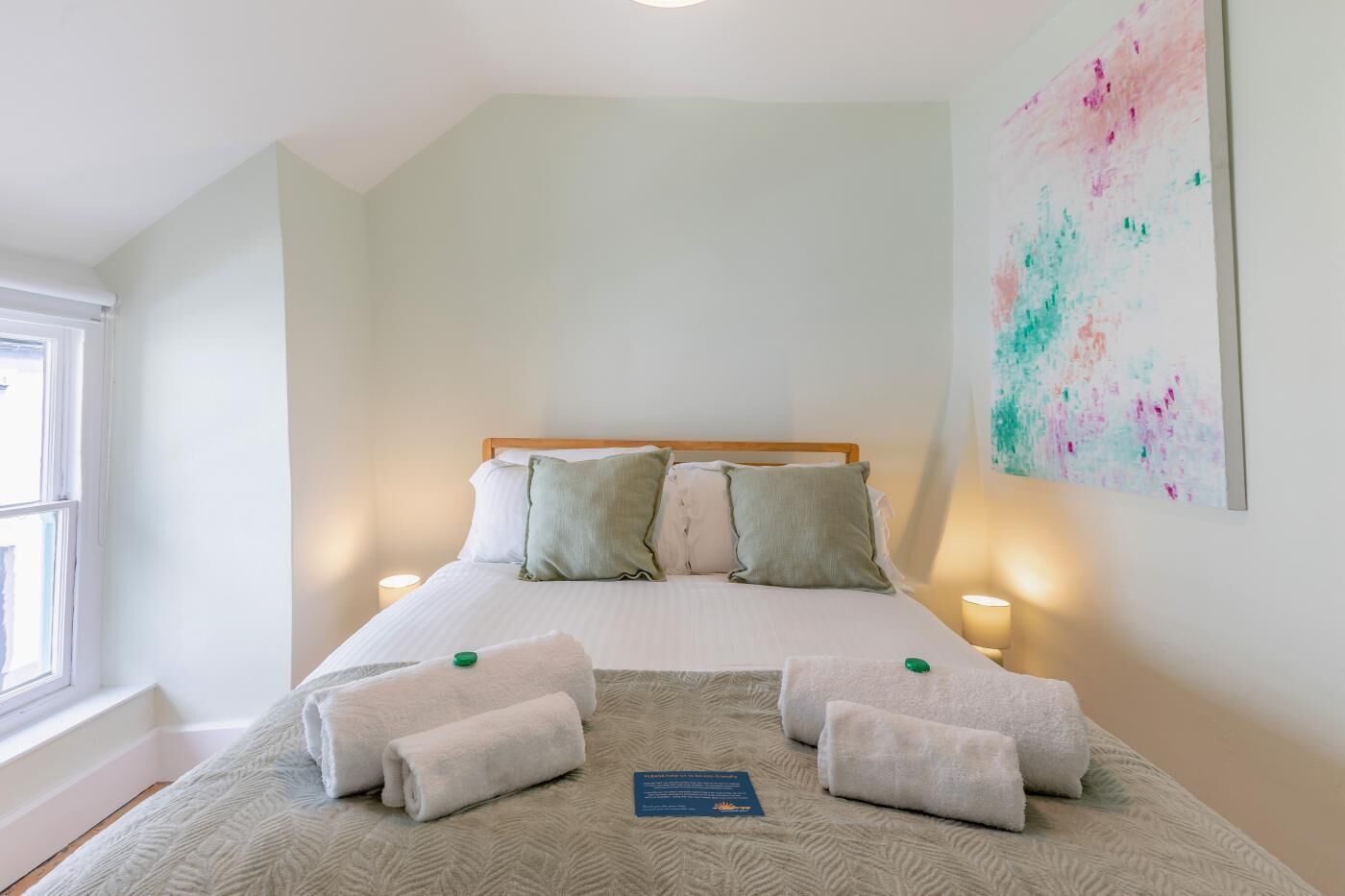 Une chambre à coucher avec un lit, des oreillers, une couverture, une serviette, une lampe de chevet et un tableau au mur.