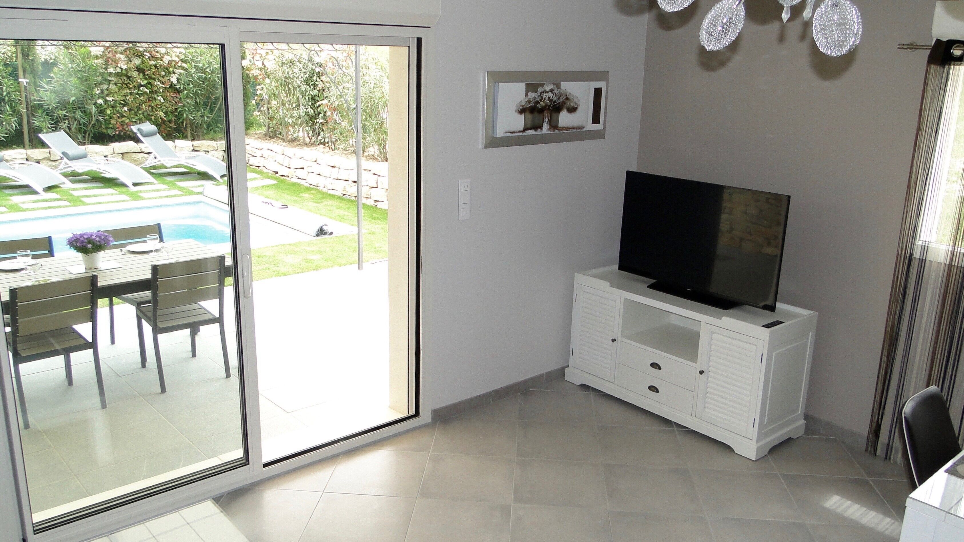 A room with a sliding glass door leading to a patio with a pool and lounge chairs, a flat-screen TV on a white cabinet, and a framed picture on the wall.