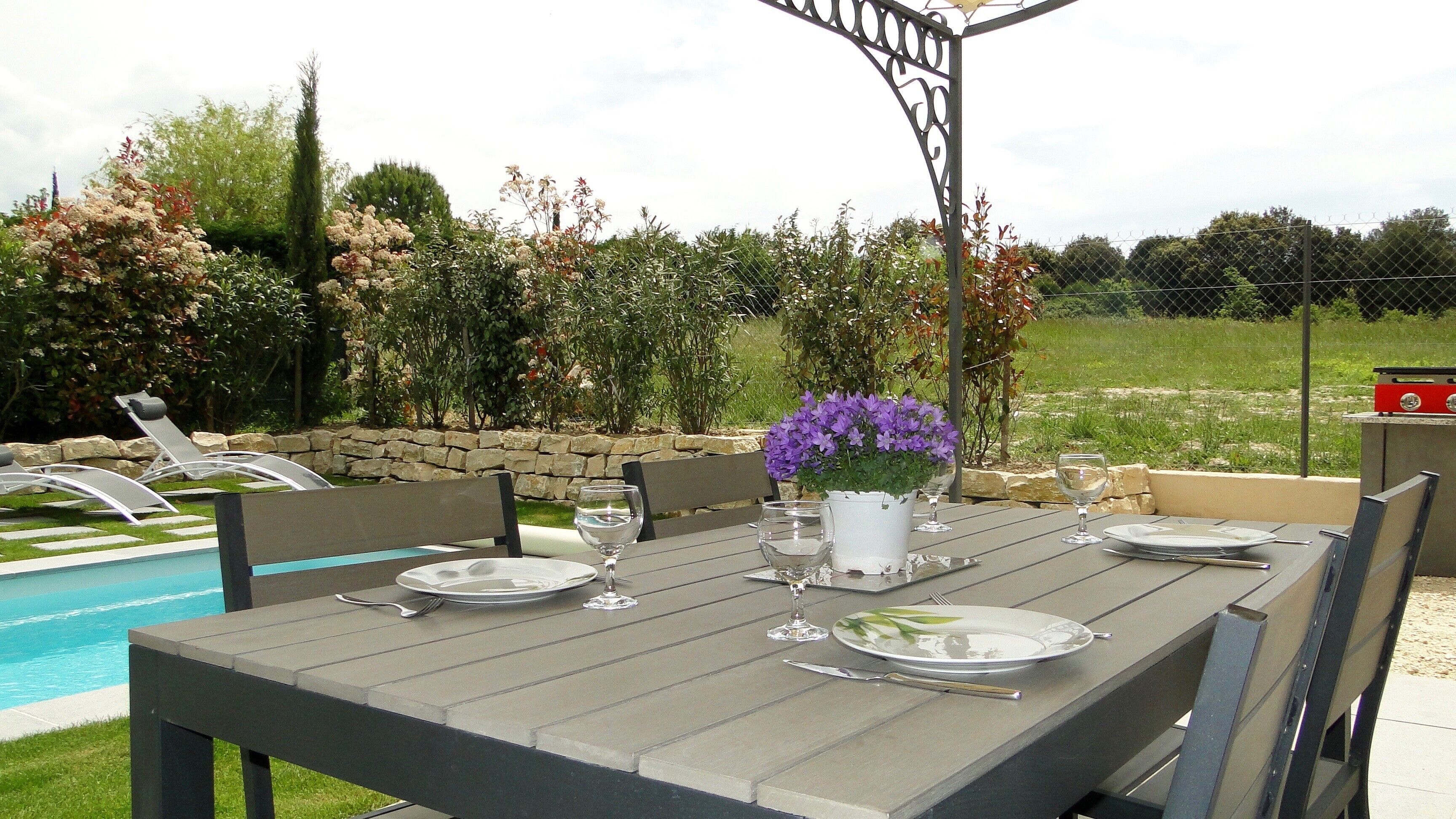 An outdoor dining area with a table set for four, surrounded by chairs, a stone wall, and a pool in the background.