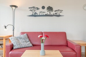 A red sofa with a patterned pillow, a vase with flowers, a wall decal of a savannah scene, a floor lamp, and a small table. - Perfekt fur Familien - Grosse Ferienwohnung mit Balkon in Sellin (Sellin)