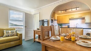A cozy room with a sofa, a dining table set for breakfast, and a kitchen area with wooden cabinets and appliances. - 2 Bed in Braunton (oc-87111) (Croyde)