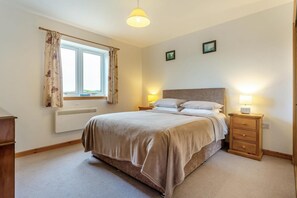 A bedroom with a bed, bedside lamps, a window with curtains, a radiator, and a wooden nightstand. - 4 Bed in Lairg (oc-ca417) (Lochinver)