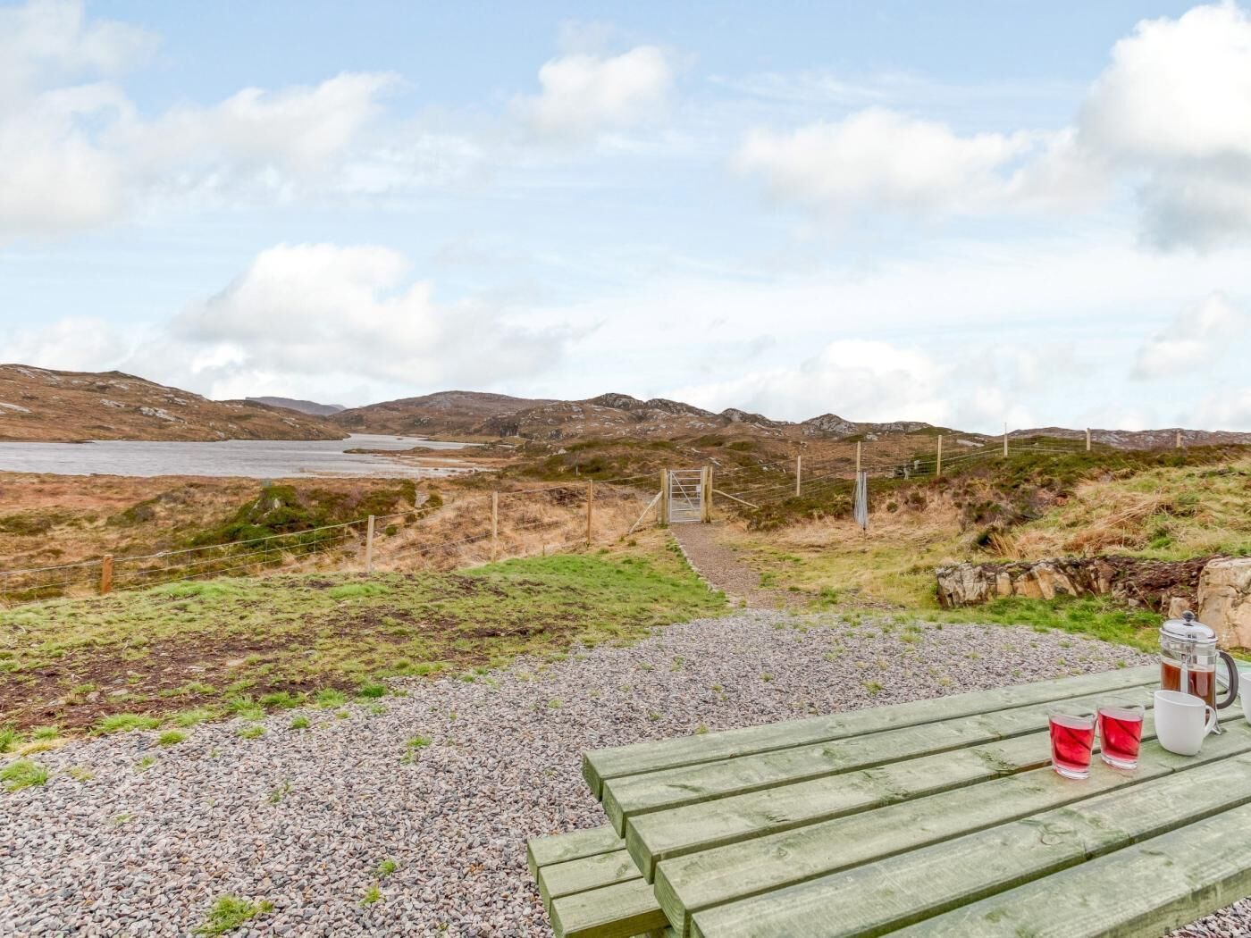 A scenic outdoor area with a gravel path leading to a wooden bench, a metal gate, and a view of a lake and hills.