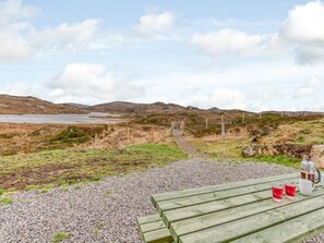 A scenic outdoor area with a gravel path leading to a wooden bench, a metal gate, and a view of a lake and hills.