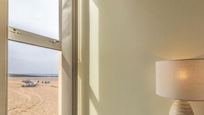 Een kamer met een raam dat uitkijkt op een strand, een lamp op een nachtkastje, en een deur met een handgreep.