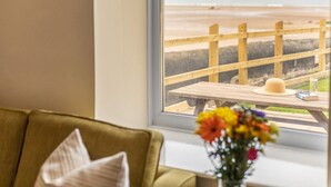 Een kamer met uitzicht op het strand, een houten tafel met een hoed en een boek, en een groene bank.