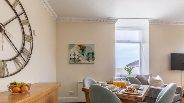 Uma área de refeições com mesa de madeira, duas cadeiras, uma cesta de frutas e um quadro na parede.