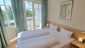Una cama doble con ropa blanca, una mesita de noche con lámpara y una ventana con cortinas.