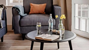 A modern living room with a dark grey sofa, a round coffee table with a book, glasses, and bottles, and a vase with yellow flowers. - 3 Bed in Braunton (oc-87754) (Croyde)