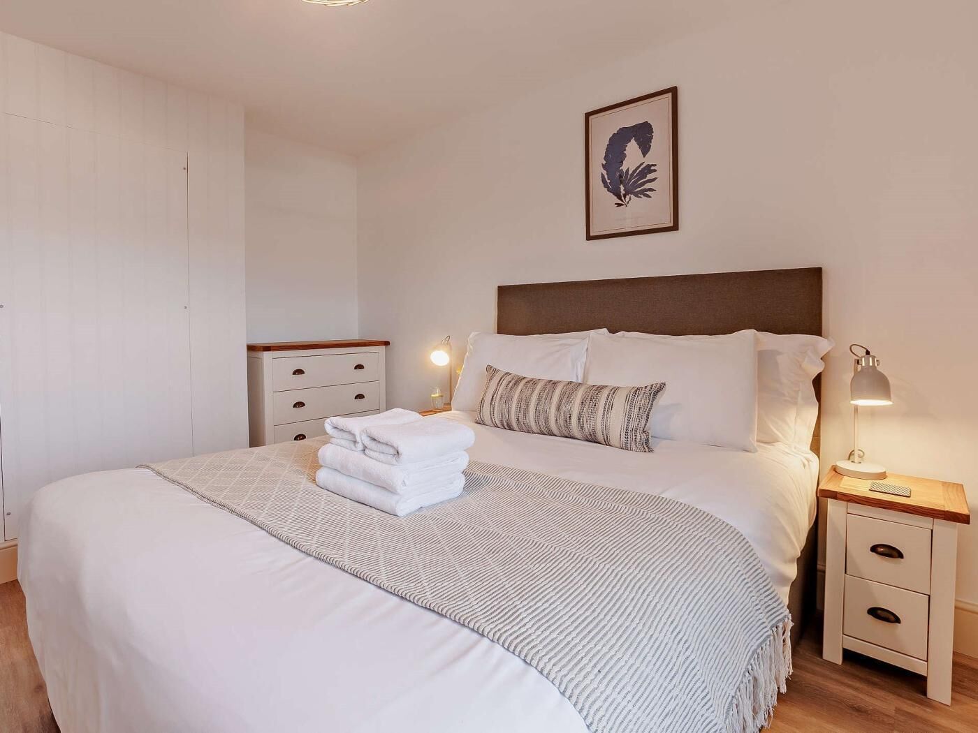 A neatly made bed with white linens and a folded towel, a bedside lamp, a nightstand with drawers, and a framed artwork on the wall.