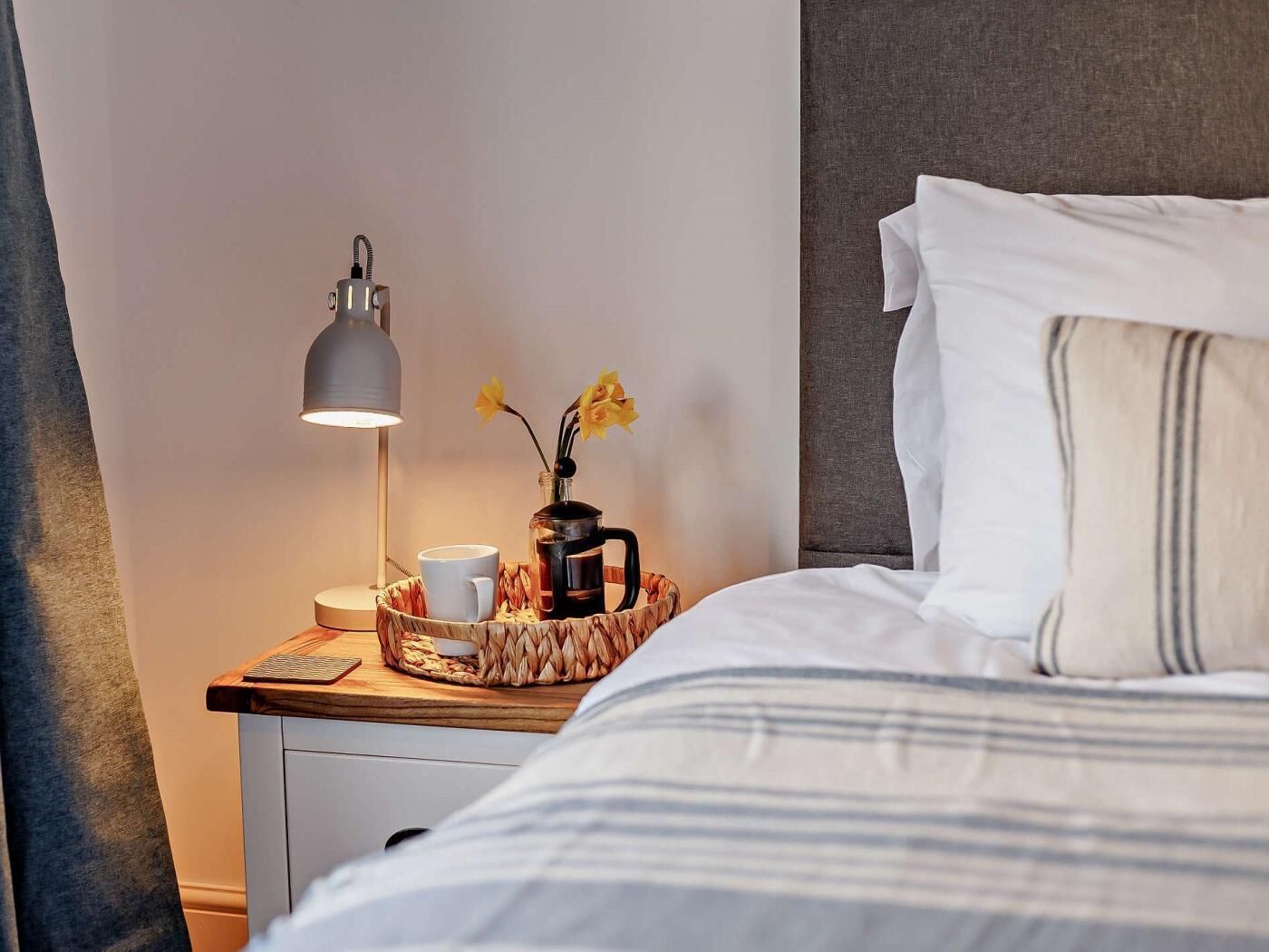 A neatly made bed with white and striped bedding, a bedside table with a lamp, a coffee pot, a cup, and a vase with flowers.