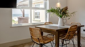 Uma sala de jantar com mesa e cadeiras, um vaso de planta e vista para o mar através de uma janela.