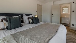 Quarto com cama, travesseiros, abajur, luminária pendente e porta de acesso a um banheiro.