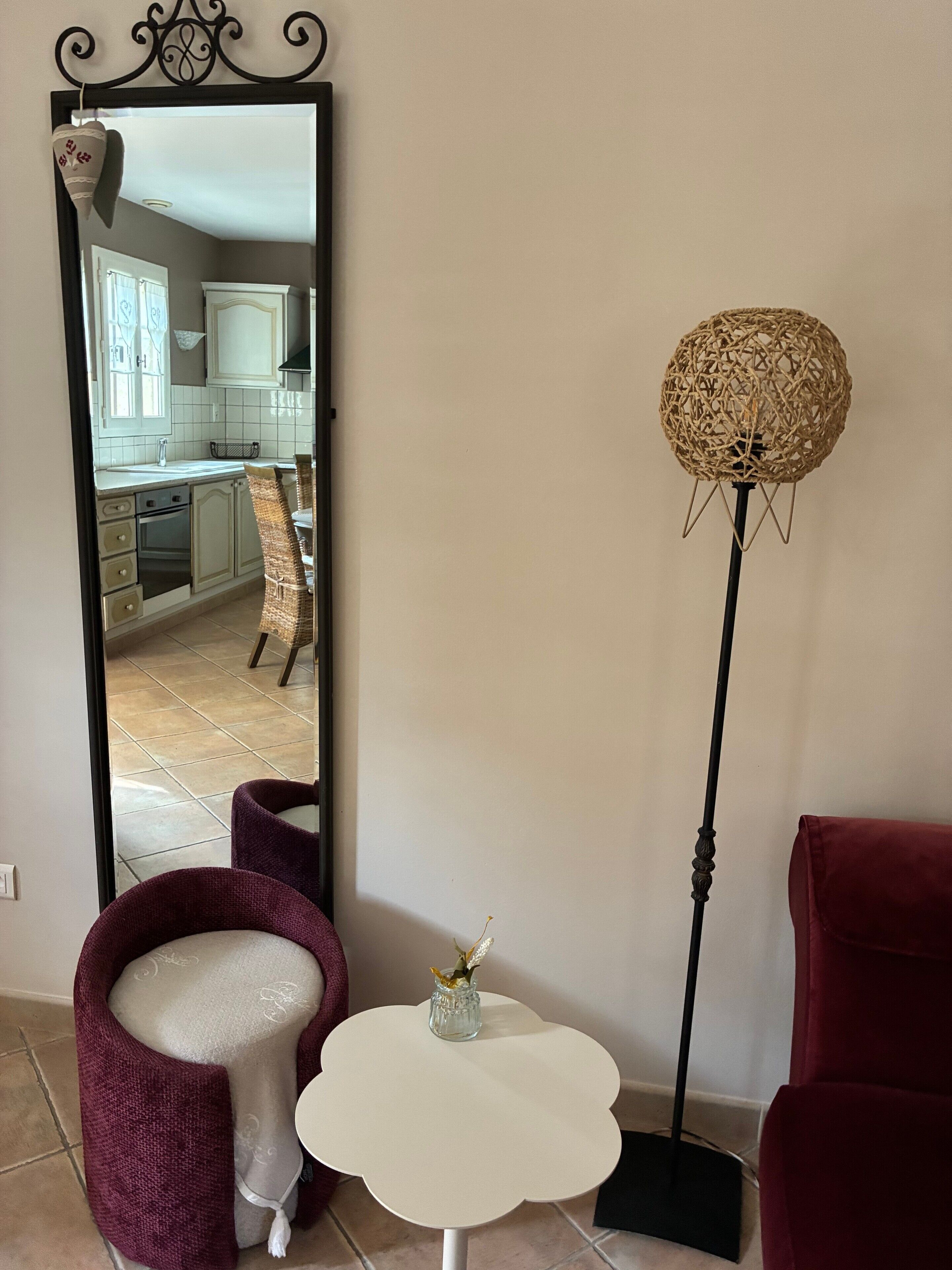 A room with a large mirror reflecting a kitchen, a round floor lamp, a small white table, and two chairs.